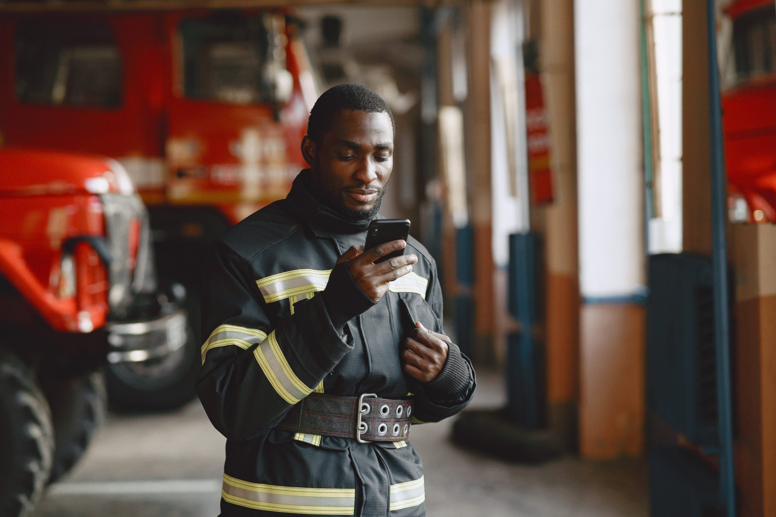 Firefighter in uniform checking his smartphone inside a fire station with fire trucks in the background.